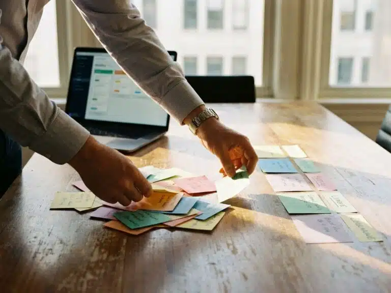 Geschäftsführer ordnet bunte Kanban-Karten auf Holztisch von chaotischem Stapel in organisierte Spalten, Laptop zeigt Workflow-Diagramme