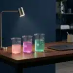 Drei Glasmessbecher mit leuchtenden rosa, magenta und türkisfarbenen Flüssigkeiten auf modernem Schreibtisch vor marineblauem Hintergrund