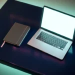 Aufgeklapptes Laptop mit leuchtendem Bildschirm neben geschlossenem Notizbuch mit rosa Lesezeichenband auf dunklem Marineblau-Schreibtisch.