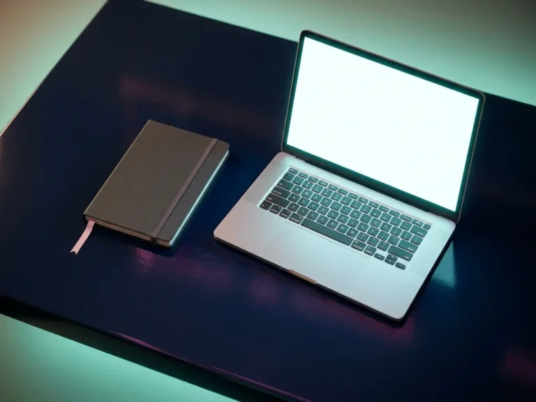 Aufgeklapptes Laptop mit leuchtendem Bildschirm neben geschlossenem Notizbuch mit rosa Lesezeichenband auf dunklem Marineblau-Schreibtisch.