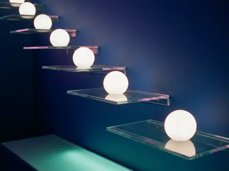 Kaskadierende Glasregale mit leuchtenden weißen Kugeln auf Marineblauem Hintergrund, rosa und türkises Lichtambiente.