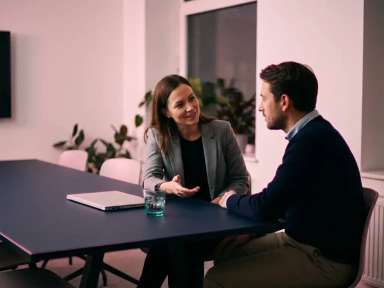Zwei Kollegen im ruhigen Gespräch an einem dunklen Konferenztisch, modernes Büro mit sanftem rosa Umgebungslicht.