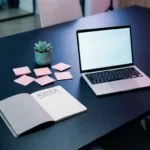 Dunkler Marineblau-Konferenztisch mit aufgeklapptem Notizbuch, schlankem Laptop, rosa Haftnotizen und einem kleinen Teal-Sukkulenten.