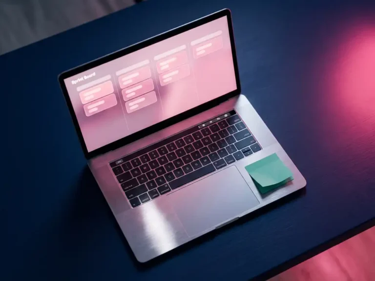 Laptop auf dunkelblauem Schreibtisch zeigt ein minimalistisches Sprint-Board mit rosafarbenen Aufgabenkarten und grünem Haftnotiz-Akzent.