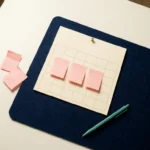 Minimalistischer Wandkalender auf dunkelblauem Untergrund mit drei rosafarbenen Haftnotizen und einem türkisfarbenen Stift, Flat-Lay-Aufnahme.