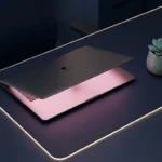 Schlankes Laptop auf dunkelblauem Schreibtisch mit rosa Bildschirmlicht und kleinem türkisfarbenen Sukkulenten daneben.