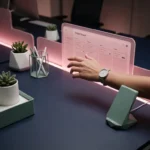 Hand eines neuen Teammitglieds greift nach einem leuchtend rosa Projektboard auf einem dunkelblauen Schreibtisch mit minimalistischen Workspace-Accessoires.