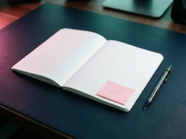 Aufgeschlagenes Notizbuch mit weißen Linien, rosa Haftnotiz mit Häkchen und mechanischem Bleistift auf dunkelblauer Oberfläche.