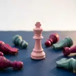 Rosa Schachkönig steht aufrecht auf dunkler Oberfläche, umgeben von gefallenen Figuren in Magenta und Türkis.