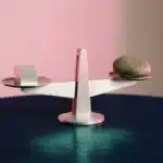 Minimalistische Waage auf dunkelblauem Untergrund, eine Seite mit geometrischem Würfel, andere mit organischem Stein, rosa Gegenlicht.