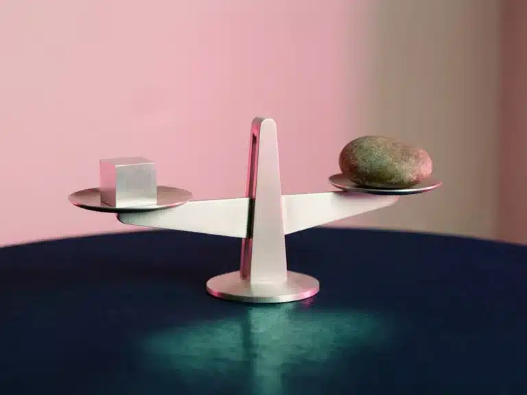 Minimalistische Waage auf dunkelblauem Untergrund, eine Seite mit geometrischem Würfel, andere mit organischem Stein, rosa Gegenlicht.